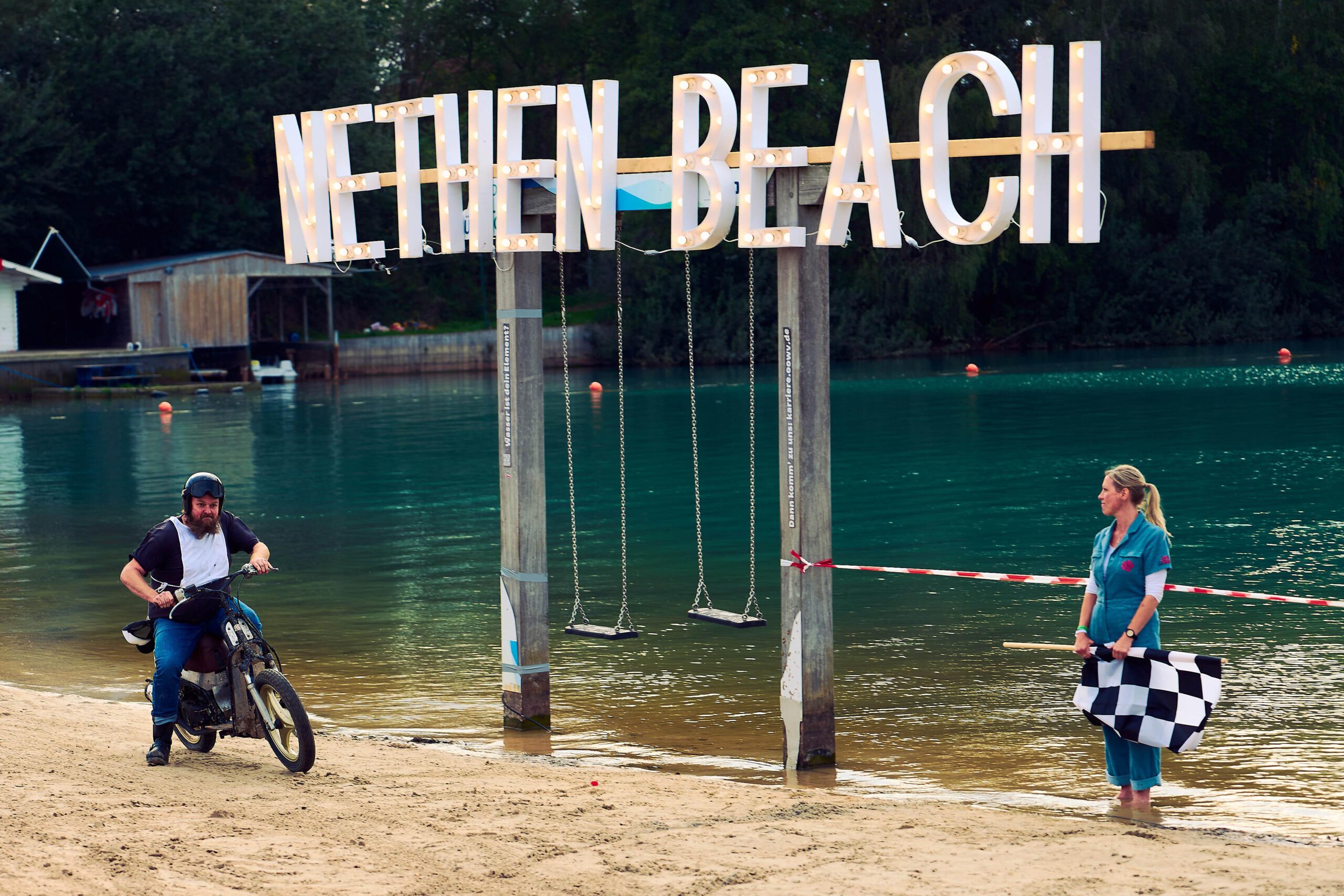 Rider on a small motorcycle at the start of the beach race under the Nethen Beach sign, a woman holding the chequered flag
